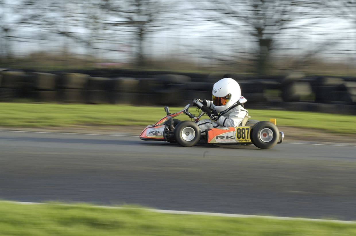 A young child in a white helmet and racing suit drives a go-kart on a paved track.